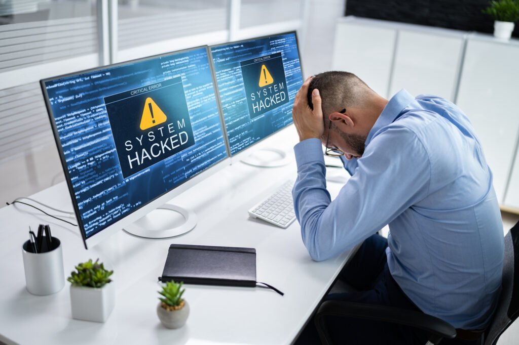 A person sits in frustration at a desk, where two monitors ominously display System Hacked warnings, highlighting the urgent need for effective penetration and security testing.