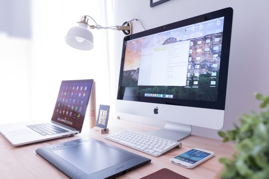 A workspace featuring an iMac and MacBook on a desk, seamlessly integrating IoT in industries, alongside a tablet, iPhone, keyboard, mouse, desk lamp, notepad, and a small plant.