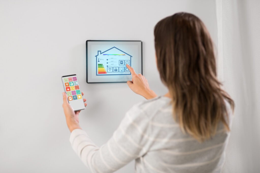 A person uses a tablet mounted on a wall to control a smart home, holding a smartphone in the other hand. Both screens display various control options for home automation, showcasing how IoT in industries seamlessly integrates into everyday living.