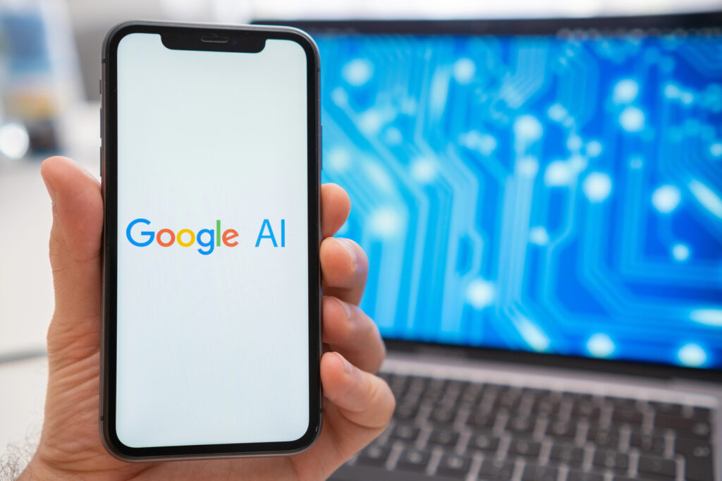 A hand holds a smartphone displaying the Google AI logo, while in the background, a blurred laptop subtly hints at the power of Google Cloud AI with its intricate circuit pattern.